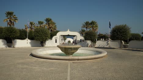 Fountain-on-the-square-at-the-entrance-to-Kallithea-Springs-and-Beach,-trees-around-and-clear-blue-sky