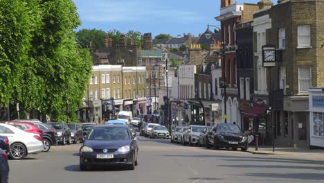 Traffic-of-Cars-Going-Up-Steep-Hill-Past-Local-Shops-in-Blackheath-Village,-South-East-London