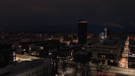 Aerial-view-low-over-night-lit-streets-and-buildings,-towards-the-Torni-hotel-and-the-railway-station,-winter-in-Tampere-city,-Finland