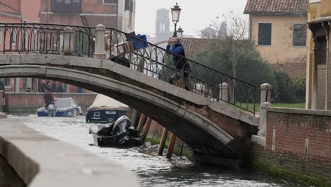 A-working-man-pushing-a-specially-designed-trolley-through-a-typical-venetian-bridge,-crossing-the-canal
