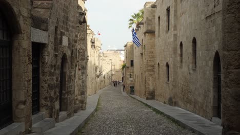 Narrow-street-of-the-Old-Town-of-Rhodes,-Tall-stone-buildings-on-both-sides-and-rocky-road