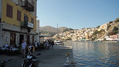 People-at-the-restaurant-and-motorbikes-passing-by-on-the-narrow-street,-Picturesque-houses-on-the-hill-in-the-background,-Harbor-of-Symi-Island