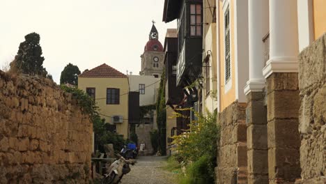 Enge-Straße-Mit-Steinmauern-Und-Traditionellen-Häusern-In-Der-Altstadt-Von-Rhodos,-Im-Hintergrund-Der-Uhrturm