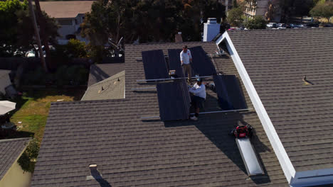Aerial-orbit-around-the-front-of-a-house-rooftop-with-workers-installing-and-performing-maintenance-on-multiple-solar-panels-in-Los-Angeles,-California---drone-shot