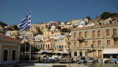 Colorful-houses-of-Ano-Symi-and-greek-flags-waving-in-the-wind-in-the-harbour