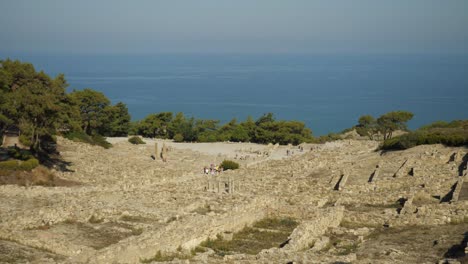 Looking-down-from-the-hill-of-Acropolis-of-Kamiros,-Tourists-walking-among-the-ruins,-Mediterranean-Sea-in-background