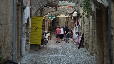 Tourists-walking-on-the-rocky-street-of-the-Old-Town-of-Rhodes,-Green-pharmacy-sign-flashing