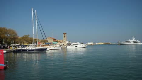 Tourboats-and-yachts-in-the-harbour-of-Rhodes,-Clocktower-of-the-Evangelist-Church-in-background
