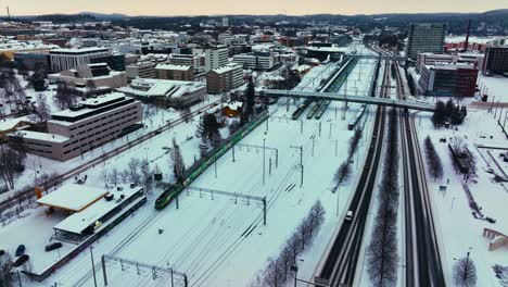 Aerial-view-following-a-train-leaving-a-snowy-railway-station-in-Jyvaskyla,-Finland---reverse,-drone-shot