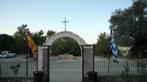The-gate-of-a-small-orthodox-church-with-a-cross-on-top-of-it,-Agios-Nikolaos-Fountoukli,-Greek-flag-and-Greek-Orthodox-Church-flag-waving