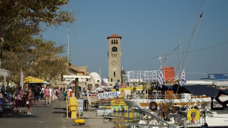 Bunter-Hafen-Von-Rhodos-Stadt,-Ausflugsboote,-Touristen-Und-Der-Glockenturm-Der-Evangelistenkirche-Im-Hintergrund