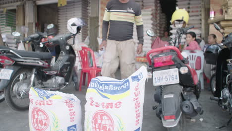 Hombre-Llevando-Una-Gran-Bolsa-De-Arroz-Fuera-De-Un-Puesto-En-El-Mercado-De-Arroz-Al-Aire-Libre-En-Camboya