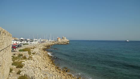 Wide-angle-view-of-the-harbour,-the-windmills-and-St-Nicholas-Fort-as-sea-waves-crashing-on-the-rocks,-Hills-of-Turkey-in-background