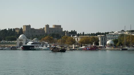 Small-pirate-tour-ship-passing-by-in-the-harbour-of-Rhodes,-The-fort-of-the-Old-Town-in-background