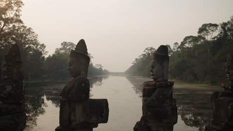 Inclínese-Hacia-Abajo-Sobre-Estatuas-De-Guerreros-Khmer-Subiendo-A-Un-Puente-Sobre-Un-Río-Tranquilo