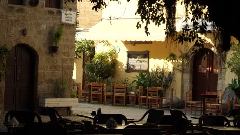 Empty-tables-of-outdoor-restaurants-in-the-Old-Town-of-Rhodes,-Cat-passing-by