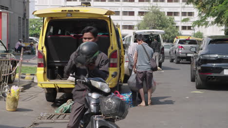 Straßenszene-In-Phnom-Penh-Mit-Zwei-Männern,-Die-Einen-Schweren-Sack-Reiskorn-Hinter-Ein-Taxi-Laden
