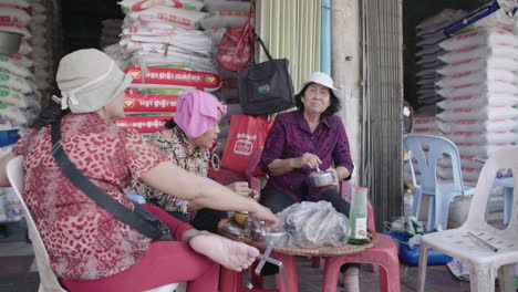 Viejas-Mujeres-Jemeres-Compartiendo-Una-Comida-Frente-A-Su-Puesto-En-El-Mercado-Al-Aire-Libre-En-Camboya