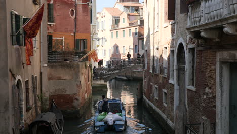 Blue-Boat-Passing-By-in-the-Canal-of-Venice,-Tourists-on-Bridge-and-Venetian-Flags-on-Buildings