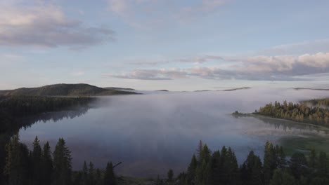 Scandinavian-forest-landscape-reflections-in-tranquil-misty-lake-below-morning-skies-DRONE