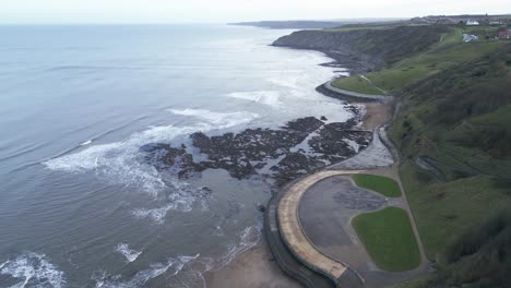 Slow-motion-aerial-shot-of-small-waves-approaching-vacant-barren-land-beside-a-hill-in-Scarborough-bay,-England
