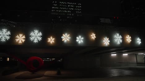 Collection-of-a-glowing-snowflake-christmas-light-decorations-cover-the-side-of-a-bridge-walkway-in-a-dimly-lit-city-at-night-with-different-sizes-and-color