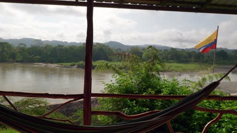 The-flag-of-Ecuador-waves-in-the-breeze-with-a-hammock-in-the-foreground-looking-out-over-a-balcony-at-the-Amazon-rainforest-across-the-Rio-Napo-in-Ecuador,-South-America