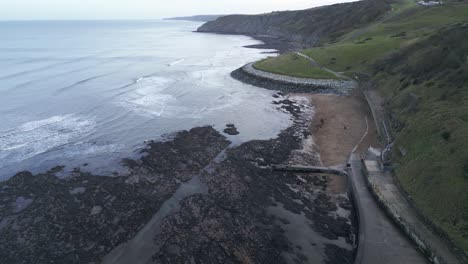 Downward-aerial-shot-of-a-vacant-river-shore-with-black-stones-on-it-and-some-waves-approaching-coast-during-evening-in-Scarborough,-England