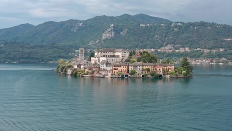 Isola-di-San-Giulio-on-Lake-Orta,-Italy,-with-historical-buildings-surrounded-by-water-and-green-hills-in-the-background,-aerial-view