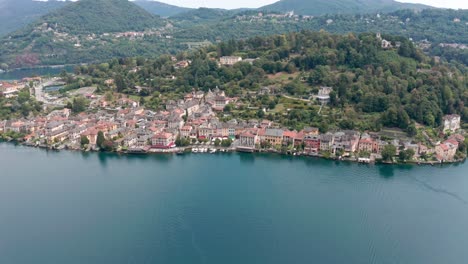 Isola-di-San-Giulio-on-calm-Lake-Orta-in-Italy,-showcasing-historic-architecture-with-overgrown-hills-in-the-background,-aerial-view