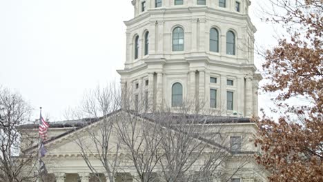Edificio-Del-Capitolio-Del-Estado-De-Kansas-Con-Banderas-Ondeando-En-Topeka,-Kansas-Con-Video-De-Plano-Medio-Inclinado-Hacia-Abajo
