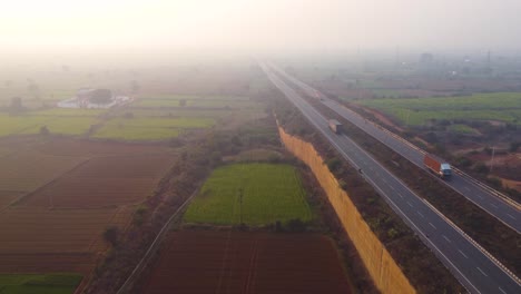 Luftdrohnenaufnahme-Von-Lastwagen,-Die-Auf-Einer-Autobahn-Durch-Bauernhöfe-Im-Ländlichen-Gwalior-In-Indien-Fahren