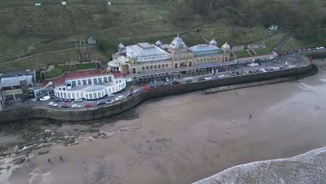 Cinematic-shot-of-Scarborough-Spa-with-cars-parked-in-front-of-it-and-a-beautiful-beach-with-small-tides-in-it-in-Scarborough,-England