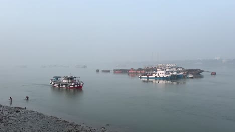 Toma-Cinematográfica-De-Un-Ferry-Navegando-Por-Un-Río-Y-Algunas-Personas-Bañándose-En-Una-Mañana-De-Invierno-En-El-Ghat-Del-Río-Hoogly-En-Calcuta,-India.