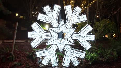 Centred-push-in-on-large-white-snowflake-christmas-decoration-sat-on-a-verge-in-a-local-city-park-in-the-Canadian-city-of-Vancouver-at-night-causing-a-festive-glow