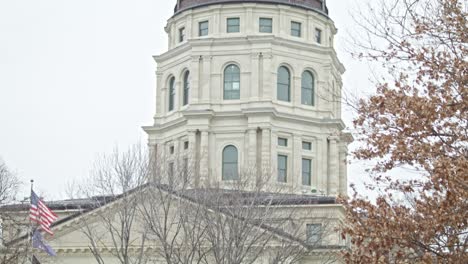 Kansas-state-capitol-building-with-flags-waving-in-Topeka,-Kansas-with-medium-shot-video-tilting-down-from-dome-in-slow-motion