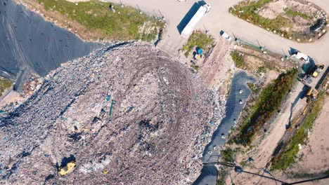 Very-high-drone-shot-above-a-mountain-of-garbage,-a-truck-is-pushing-the-garbage,-there-are-lots-of-birds-flying-over-the-trucks