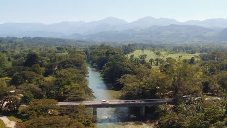 White-overland-vehicle-with-rooftop-tent-crossing-a-bridge-over-shallow-river-cutting-through-woodland-and-tree-lined-banks