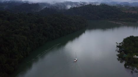 High-angle-aerial-overview-of-sailboat-going-up-river-in-misty-tropical-forest