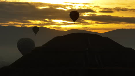 Telephoto-drone-shot-of-a-silhouette-pyramid-and-hot-air-balloons,-sunrise-in-Mexico
