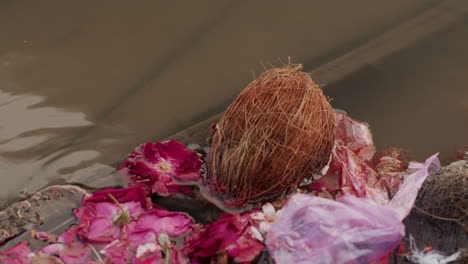 Close-up-of-offering-flowers-and-coconut-mixed-with-plastic-bag-floating-in-river