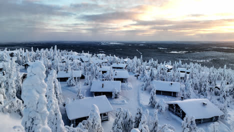 Flying-over-snowy-trees,-toward-a-cottage-village-on-top-a-fell,-winter-sunset-in-Lapland---Aerial-view