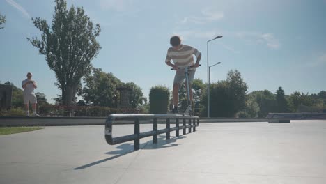 Man-on-stunt-scooter-perform-slide-on-skatepark-metal-handrail,-sunny-summer-day