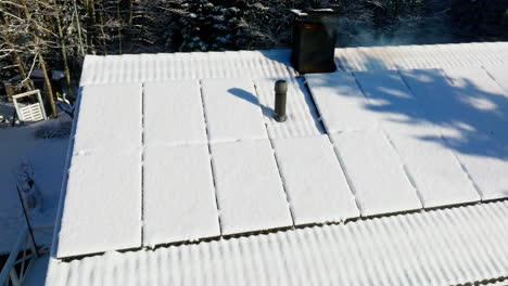 Drone-circling-sunlit,-snow-covered-solar-cells-on-a-house-with-a-smoking-chimney