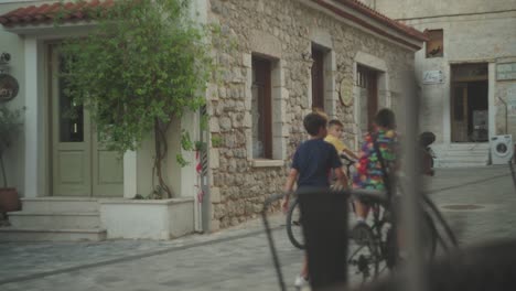 Una-Mujer-Que-Viaja-Camina-Por-La-Calle-Mientras-Los-Niños-Andan-En-Bicicleta-Frente-A-Un-Antiguo-Edificio-De-Ladrillo-En-Una-Ciudad-Mediterránea