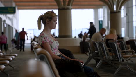 Young-woman-sitting-alone-in-airport-terminal-using-laptop-with-passengers-walking-in-background