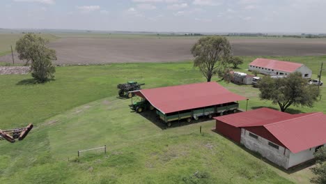 4K-Luftaufnahmen-Eines-Bauernhauses-Mit-Landwirtschaftlichen-Geräten,-Einer-Hochgeschwindigkeitsspritze-Für-Feldfrüchte,-Die-Um-Den-Bauernhof-Herumfährt