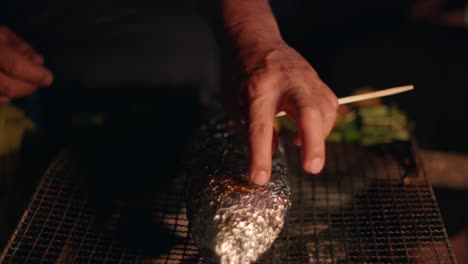 Aluminum-foil-wrapped-fish-sitting-on-barbecue-grill-to-cook-being-checked-by-hand-for-tenderness,-filmed-as-closeup-slow-motion-shot