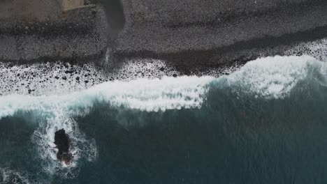 Aerial-view-of-waves-crushing-into-a-pebble-beach,-turquoise-water-and-white-foam