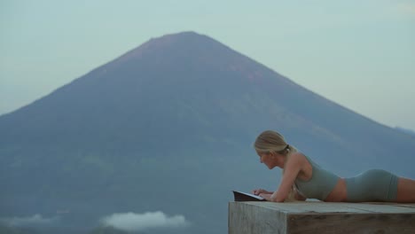 Woman-laying-on-platform-while-writing-in-journal-with-mount-Agung,-self-care-concept